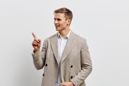 Confident young man in stylish beige blazer and white shirt pointing with finger while smiling, standing against plain white background. Studio portrait, fashion, success, communication, professional, casual style, positive expression.の写真素材