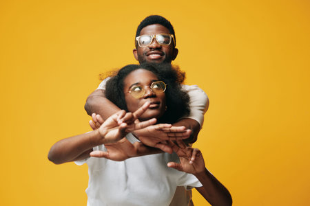 Young African couple embracing on a vibrant yellow background, showcasing joy and connection with stylish eyewear and casual clothingの写真素材
