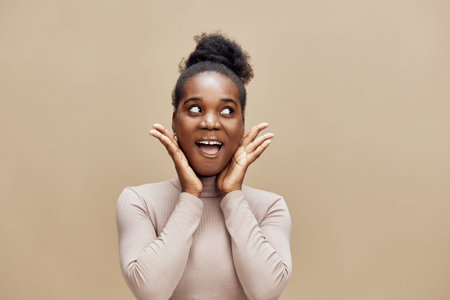 Happy young African woman expressing joy with hands on her cheeks, against a warm beige background, perfect for wellness and positivity conceptsの写真素材