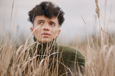 Candid portrait of a young person peering through tall dried grass, conveying authenticity,...