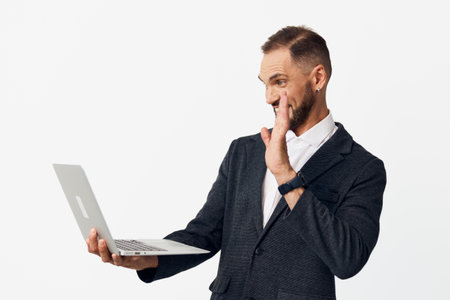 A professional man stands on a solid colored background, holding a laptop and showing thoughtful, confident emotion as he engages with the screen.の写真素材
