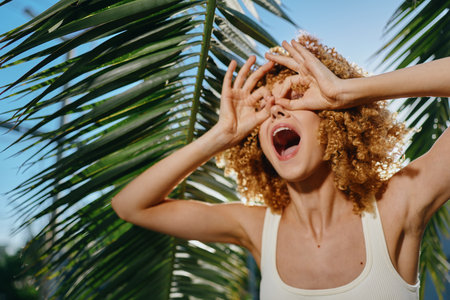 young woman with curly hair posing joyfully near palm leaves, wearing white tank top and expressing summer happiness outdoors under bright sunlightの写真素材