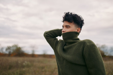Young man in a green turtleneck sweater stands contemplative in an open field, face partly hidden,...