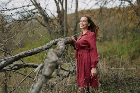 An authentic portrait of a woman in a deep maroon dress posing beside a weathered log amid autumn brush, conveying genuine spirit and grounded, believable mood.の写真素材
