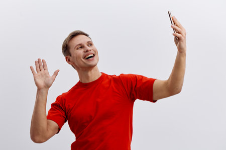 Smiling young man taking selfie with mobile phone, waving hand, wearing bright red t-shirt, happy expression, isolated on plain white background, casual style, lifestyle concept, cheerful mood.の写真素材