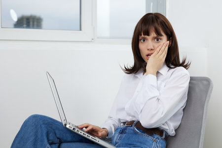 A surprised young woman sits in a bright office with a laptop on her lap, covering her mouth with...