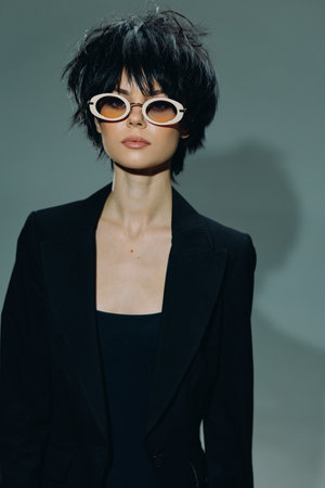 Stylish woman with short black hair and unique white sunglasses wearing a black blazer against gray backgroundの写真素材