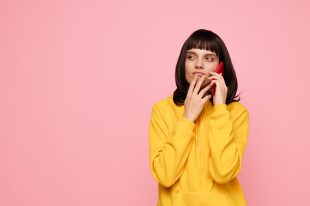 A young woman wearing a bright yellow hoodie talks on a red mobile phone against a soft pink backdrop, creating a thoughtful, focused mood in a clean, well lit studio environment.の写真素材