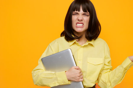 A frustrated young woman stands against a bright orange backdrop, clutching a laptop and showing signs of stress, confusion, and strain while thinking about a tough task or deadline.の写真素材