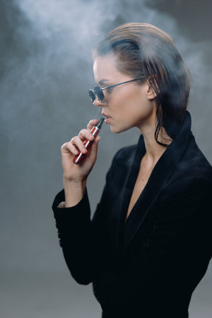 Stylish woman vaping with smoke in dark background, wearing sunglasses and black blazer, modern vapor lifestyle concept.の写真素材