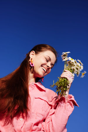 Young woman smiling brightly while holding a bouquet of daisies against a clear blue sky, dressed in a pink shirt, exuding joy and a sense of springの写真素材