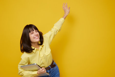 A cheerful woman with dark hair wearing a yellow shirt smiles against a bright yellow backdrop, holding a notebook and raising a hand in a joyful pose.の写真素材
