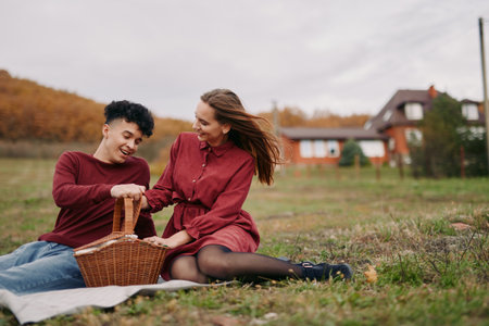 A genuine candid picnic scene of a young couple sharing laughter as they reach into a wicker basket on a blanket in an autumn meadow, capturing natural connection, trust and authenticity.の写真素材