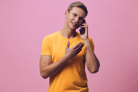 Man talking on a mobile phone smiling cheerful casual young adult wearing bright yellow t-shirt isolated on pink background studio portrait. People lifestyle conceptの写真素材
