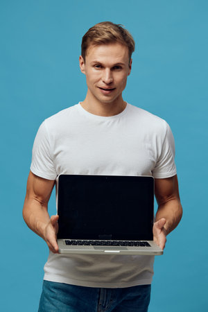 Smiling young man holding laptop computer in hands looking at camera studio portrait isolated on blue background portraying modern technology and confident mood. People lifestyle concept.の写真素材