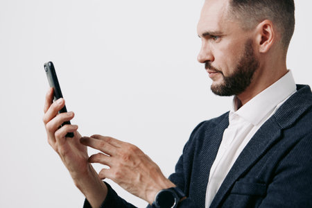 A focused business professional man stands against an isolated colored background, examining a smartphone with calm determination, confident posture, and thoughtful expression.の写真素材