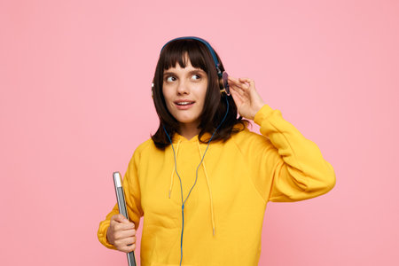 Young woman wearing a bright yellow hoodie listens to music with headphones, holding a notebook and a confident smile against a pink background, presenting a fresh, vibrant mood for everydayの写真素材
