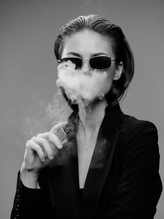 Black and white portrait of a woman vaping with sunglasses and a dark jacket in studio lighting.の写真素材