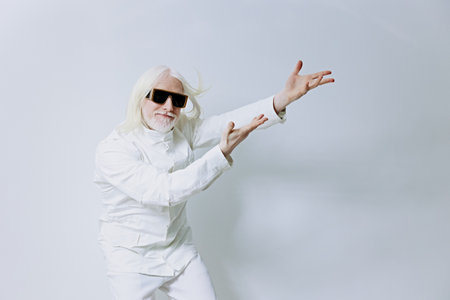 Stylish elderly man with sunglasses gesturing in a minimalist fashion studio, dressed in a white outfit against a neutral background, exuding confidence and creativityの写真素材