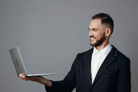 A professional man in business attire holds a laptop on an isolated colored background, exuding confidence, focus, and poised energy during a moment of digital work.の写真素材