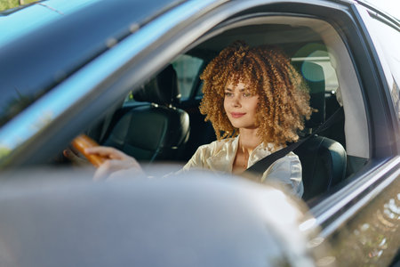 Young woman with curly hair driving a car wearing seatbelt, focused and calm, sunlight illuminating...