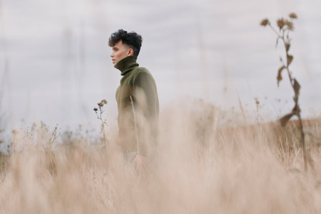 A contemplative young person in a textured green sweater walks through tall dry grass, captured with natural light and an honest perspective, emphasizing emotional authenticity and trustworthiness.の写真素材
