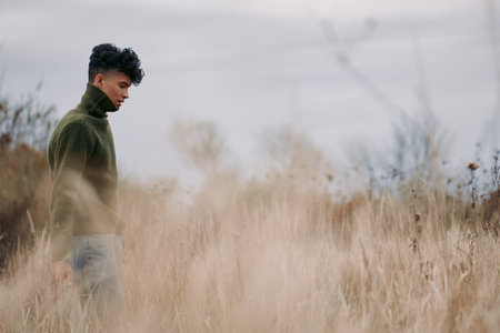 A contemplative young man walks through tall dry grass in muted overcast light, evoking authenticity and truthful emotion with natural posture, simple clothing and quiet presence.の写真素材