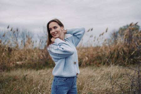 Portrait of a young woman in a cozy blue knit sweater standing in a windswept meadow, candid smile and relaxed posture conveying authentic emotion and believable everyday charm.の写真素材