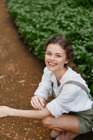Happy young Caucasian woman enjoying nature while kneeling in a green garden, wearing casual clothing and smiling brightly, embodying joy and tranquilityの写真素材