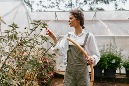 Young woman in greenhouse, wearing light overalls and a white shirt, showing joy while gardening, surrounded by lush plants, autumn colors, and natural lightの写真素材