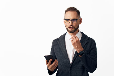A confident business professional stands against a plain background, holding a smartphone and...