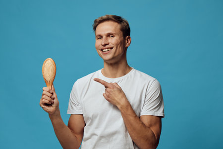 Smiling man holding hairbrush and pointing at it isolated on blue background studio portrait, casual clothing, cheerful mood, personal care concept, haircut, grooming, grooming tools, male hygiene, wellness, lifestyleの写真素材