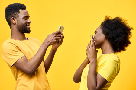 Smiling African couple taking selfies together against a vibrant yellow background, expressing joy and connection in matching yellow outfitsの写真素材