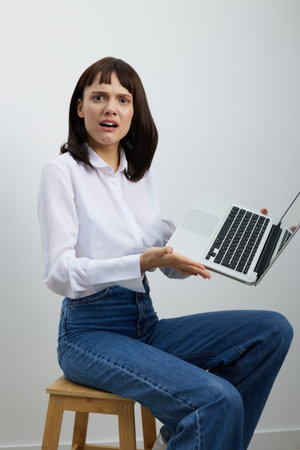 A surprised woman sits on a wooden stool, presenting an open laptop toward the camera. She wears a...
