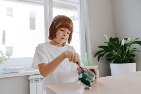 A focused boy is using a screwdriver to work on a small toy. The bright space features plants,...