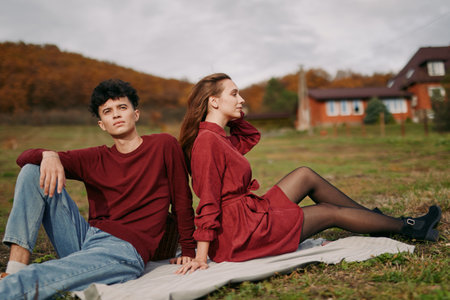 A young couple sits back to back on a blanket in an autumn field, a candid, natural portrait capturing quiet emotion, authenticity and understated credibility in a real moment.の写真素材