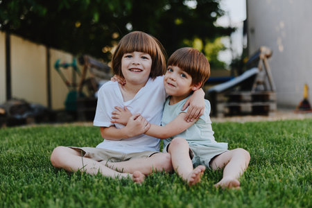 Two joyful boys sitting together on green grass, enjoying a sunny day outdoors with smiles and affectionate hugs that convey warmth and friendship.の写真素材