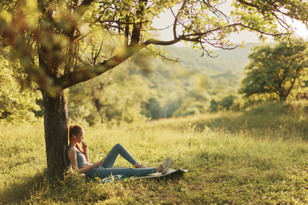 Young woman relaxing under a tree in a green field, enjoying nature and tranquility during a sunny day, evoking feelings of peace and serenityの写真素材