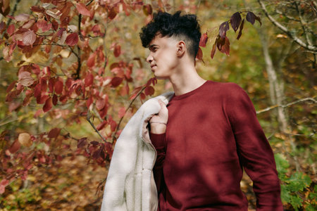 A young man stands in a autumn forest, wearing a burgundy sweater and draped jacket, conveying authenticity and credibility through a candid, natural pose.の写真素材