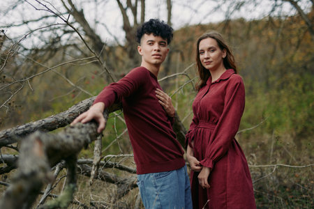 Authentic, candid portrait of a young couple outdoors, wearing burgundy outfits, standing by a rustic fence amid bare branches, conveying real emotion and genuine connection.の写真素材