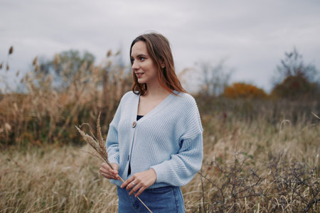 A candid portrait of a thoughtful woman in a soft blue knit cardigan standing in a sunlit field, conveying authenticity and natural charm.の写真素材