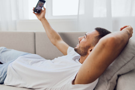 Relaxed man enjoying a video call while lounging at home, dressed casually in a cozy white t-shirt. Bright and inviting atmosphere enhances the connection.の写真素材