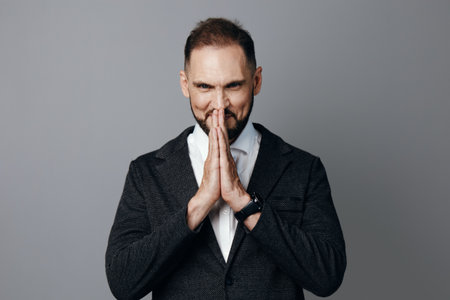 A professional man in business attire stands against a solid colored background, projecting focus and determination as hands are pressed together in a thoughtful pose.の写真素材