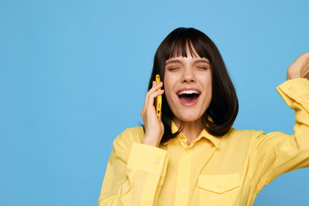 Bright portrait of a cheerful woman in a yellow shirt, smiling while talking on a mobile phone against a blue studio backdrop, conveying energetic communication, friendly mood, and modern daily life.の写真素材