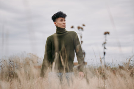 An authentic portrait of a young man standing in tall grass, wearing a dark green turtleneck, conveying a calm, grounded presence in a wide field.の写真素材