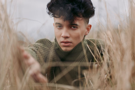 A close, intimate portrait of a young person reaching through tall dry grass, raw expression and...