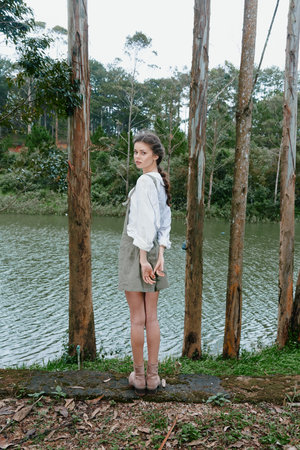 Young woman in a forest standing by a serene lake wearing a light blouse and skirt, showcasing a calm and peaceful demeanor influenced by natureの写真素材