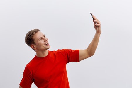 Smiling young man taking a selfie with smartphone, casual clothing, red t-shirt, cheerful expression, white background, outdoor activity, lifestyle conceptの写真素材