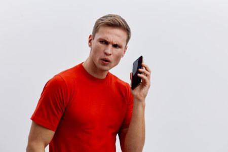 confused man holding black mobile phone in red t-shirt looking side isolated on white background cyber security conceptの写真素材