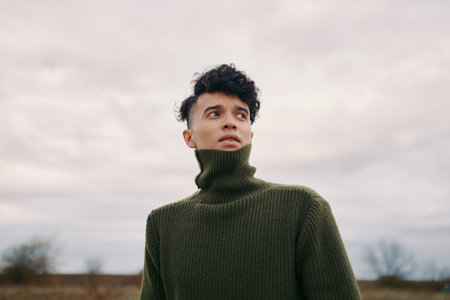 A candid portrait of a young man in a dark green turtleneck sweater, conveying authenticity and credibility through natural expression and a calm, thoughtful gaze against a muted sky.の写真素材